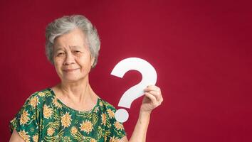 Senior woman holding a white question mark symbol with a smile while standing on a red background photo