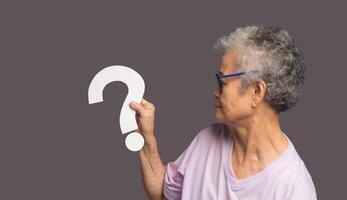 Senior woman holding a white question mark symbol with a smile while standing on a gray background photo