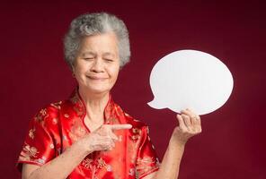 Senior woman wearing a traditional cheongsam qipao holding and pointing to a blank speech bubble and smiling while standing on a red background photo