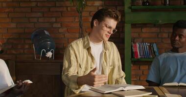 Three men sitting at a table with books and a man giving a thumbs up video