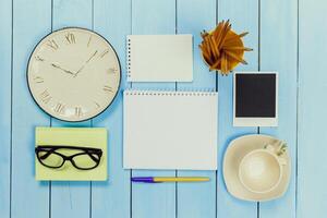 Arrangement of office supplies including a clock, notebook, and glasses on a blue wooden table in a workspace setting photo