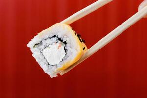 Sushi roll with cream cheese and crab held by chopsticks in front of a red background photo