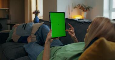 ein Frau ist Sitzung auf ein Couch mit ein Tablette Computer mit ein Grün Bildschirm video