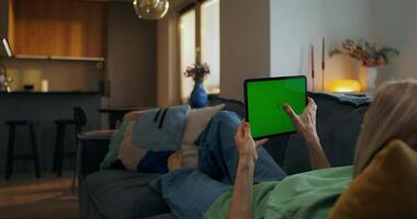 A woman is sitting on a couch with a tablet computer with a green screen video