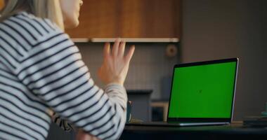A woman is sitting at a table with a laptop that has a green screen video