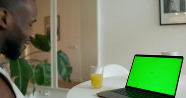 A man is sitting at a table with a laptop that has a green screen video