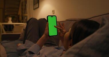 A woman is sitting on a couch and using a smartphone with a green screen at night video
