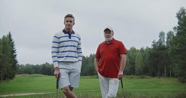 Two men of different ages are leaning on golf clubs on the golf course and smiling while looking at the camera. Dynamic camera movement in a circle video