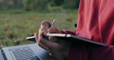 A person is writing on a notebook while using a laptop video