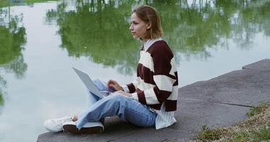 A woman sitting on the edge of a lake with a laptop video