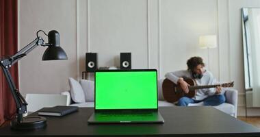 A laptop with a green screen on a table in front of a man playing guitar video