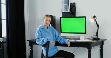 A woman in a blue sweatshirt sitting at a desk with a green screen video