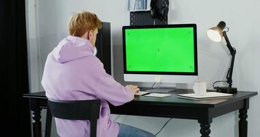 A man in a hoodie is sitting at a desk with a green screen video