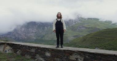 A woman standing on a stone wall in front of a mountain video