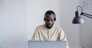 A man wearing headphones sits at a desk with a laptop video