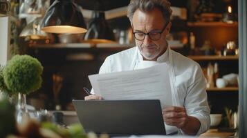 Chef reads a document on a laptop. Man in kitchen looks at paper notes. Professional works using a computer. photo
