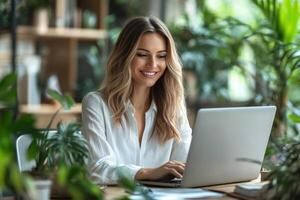 Woman at desk uses laptop. Smiling person works with computer near green plants. Focus on screen and typing hands. Natural light office. photo