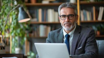 Man with computer looks at camera. Grey hair. Bookshelves in the background. Lamp and plants. Office setting. Professional man at desk. Working man portrait. photo