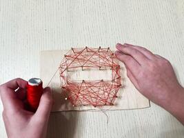 Hands crafting geometric string art with red threads on a wooden board photo