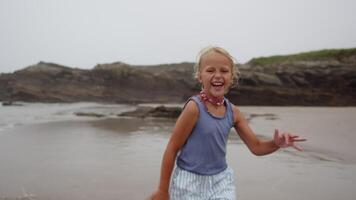 Young girl happily running towards the camera on a sandy beach with a rocky coastline in the background, filmed in slow motion video