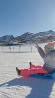 Happy child enjoying winter fun, sliding down a snowy slope on a plastic sled, surrounded by a beautiful mountain panorama under a clear blue sky video