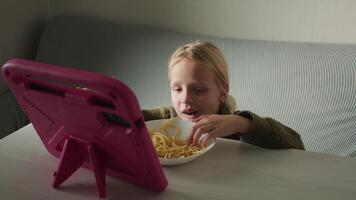 Young girl consuming spaghetti while deeply concentrated on digital tablet screen, representing contemporary digital interaction during casual mealtime experience in home environment video