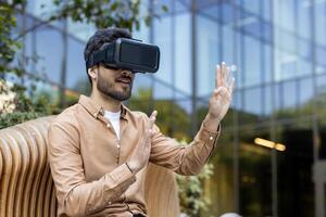 Man wearing VR headset sitting on bench, immersed in virtual reality outdoors. Expressive gestures indicate engagement with digital experience. Setting suggests innovative technology photo