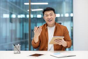Asian man in a call, sitting at his desk with a notebook. He is smiling and looks like he is enjoying the conversation photo