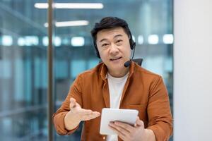 Asian man wearing a headset and holding a tablet while working in a call center, providing support to customers remotely photo