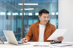 Successful joyful businessman investor doing paperwork, asian man working with documents reports and contracts. Man in casual clothes using laptop at work inside office. photo