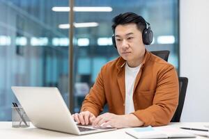 Serious confident and focused Asian man working inside office with laptop, wearing headphones, male programmer developer at workplace, coding new software. photo
