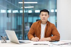 Asian businessman is in his office, concentrating and writing on a document while looking at the camera with a determined expression photo