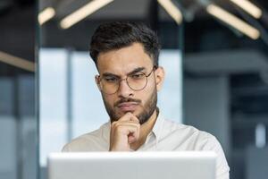 Man wearing glasses concentrating on computer screen in office setting. Serious expression suggests deep thought or analysis. Modern work environment. Keywords. office, focus, professional. photo