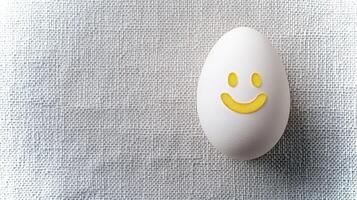 A white egg adorned with a bright yellow smiley face rests on a light-colored textured surface, bringing a playful and cheerful touch to the setting. photo