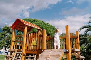 Little girl stands on a wooden slide on a playground, leaning on a fence. Back view photo