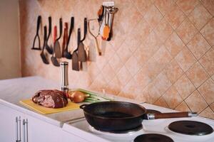 Frying pan stands on the stove in the kitchen next to raw meat and vegetables on the table photo