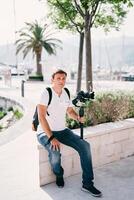Photographer with a camera on a tripod sits on a stone fence near a tree photo