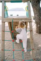 Little girl climbs a rope ladder onto a slide on a beach playground. Back view photo