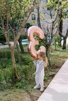 Little girl with an inflatable number 3 stands in a green garden near a path and looks at her reflection in it photo