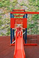 Little girl slides down a slide on a playground near a stone wall covered with ivy photo