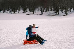 Dad with a small child sits on a sled on a snowy plain and points towards the forest. Back view photo