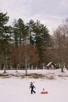 Mom is carrying a small child in a sled past a wooded hill with a stone shed. Side view photo