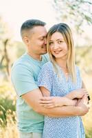 Guy hugs smiling girl from behind, touching her cheek with his nose while standing in the garden photo
