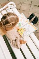 Little girl with a soft toy reads a colorful book while sitting on a white bench. Top view photo
