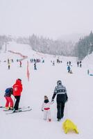 Mothers with children on skis and sleds climb a snow-covered ski slope along the red fence. Back view photo