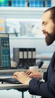 Vertical Photovoltaics factory worker monitors system performance, drinking coffee. Solar panels manufacturing plant employee enjoys beverage while using PC to track energy production and efficiency, camera B video