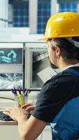 Vertical Photovoltaics factory engineer using PC to test robot arms placing solar panels on assembly lines. Professional sitting at computer desk inspecting solar plant heavy machinery units, camera B video