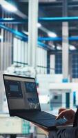 Vertical Man in photovoltaics factory monitoring system performance on laptop. Solar panels manufacturing plant employee using notebook to track energy production and efficiency, camera A close up video