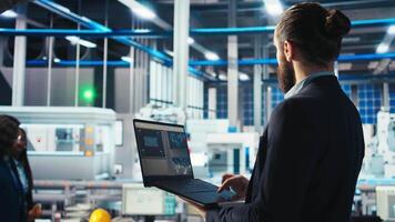 Man in photovoltaics factory monitoring system performance on laptop. Solar panels manufacturing plant employee using notebook to track energy production and efficiency, camera A close up video