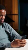 Vertical African american person takes a break from work to answer texts at home, using his smartphone to scroll social media apps. Young guy enjoying remote job at home, listens to music on phone. Camera B. video